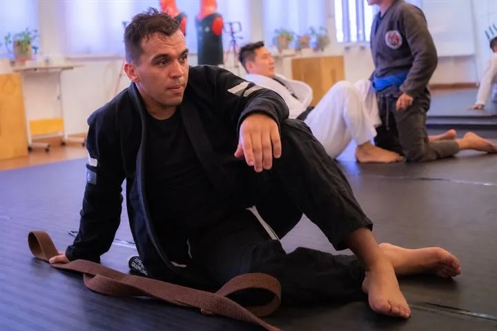 A Jeet Academy KL instructor in a black gi resting on the mats with a brown belt beside him during a grappling class